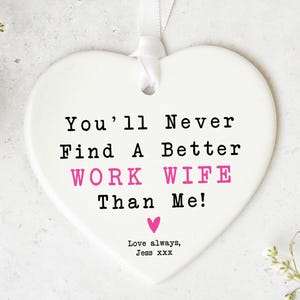 May include: A white heart-shaped ceramic plaque with the text "You'll Never Find A Better WORK WIFE Than Me!" in black and pink. Includes a pink heart and the words "Love always, Jess xxx". A white ribbon is attached.