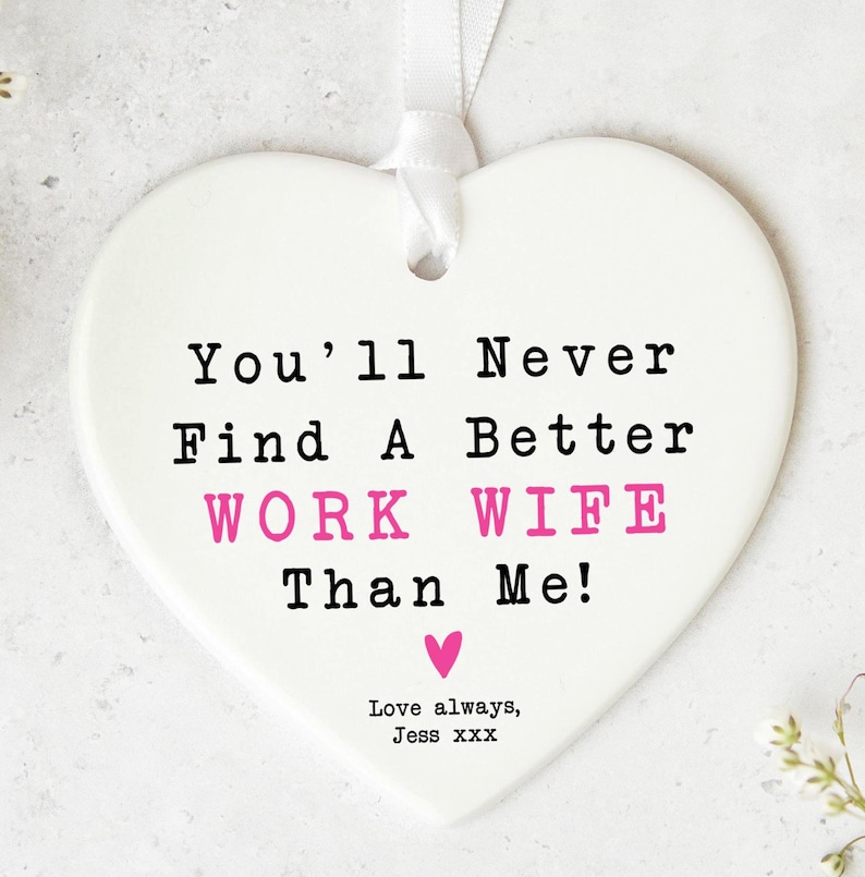 May include: A white heart-shaped ceramic plaque with the text "You'll Never Find A Better WORK WIFE Than Me!" in black and pink. Includes a pink heart and the words "Love always, Jess xxx". A white ribbon is attached.