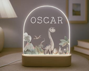 May include: A wooden night light with a clear acrylic panel featuring a dinosaur, a volcano, and a palm tree. The name "OSCAR" is printed on the panel.