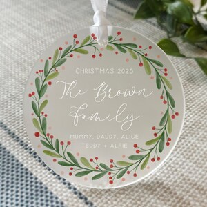May include: A round, off-white Christmas ornament with a watercolor wreath of green leaves and red berries. The text "CHRISTMAS 2025" and "The Brown Family" are written in elegant script. Additional names are listed below.
