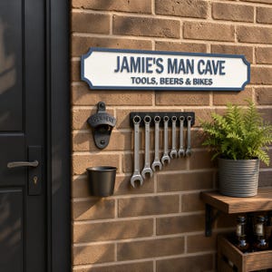 a brick wall with a sign that reads "JAMIE'S MAN CAVE" and a row of tools hanging on it. There is also a potted plant and a beer bottle on the wall.