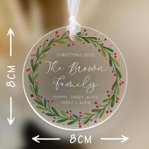 May include: A clear, round Christmas ornament, 8 cm in diameter, with a white ribbon for hanging. The ornament features a green and red wreath design around the edge. Text includes "Christmas 2025" and "The Brown Family".