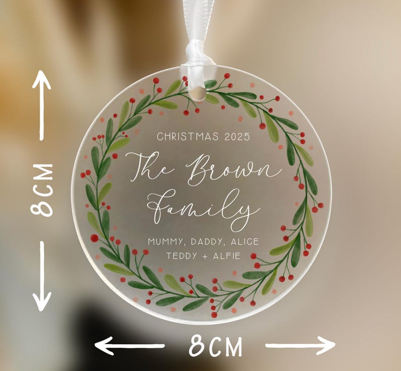 May include: A clear, round Christmas ornament, 8 cm in diameter, with a white ribbon for hanging. The ornament features a green and red wreath design around the edge. Text includes "Christmas 2025" and "The Brown Family".