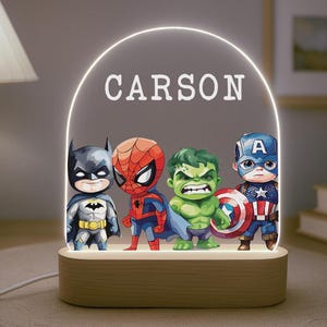 May include: A wooden night light with four superhero figures: Batman, Spider-Man, Hulk, and Captain America. The name "CARSON" is illuminated above the figures. The light emits a warm glow.