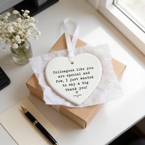a heart-shaped ceramic tag with a message, placed on a desk next to a vase of white flowers and a pen.