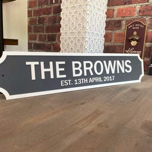 May include: A gray and white sign with the text "THE BROWNS EST. 13TH APRIL 2017". The sign has a white border and is displayed on a wooden surface. The background includes a brick wall and decorative items.