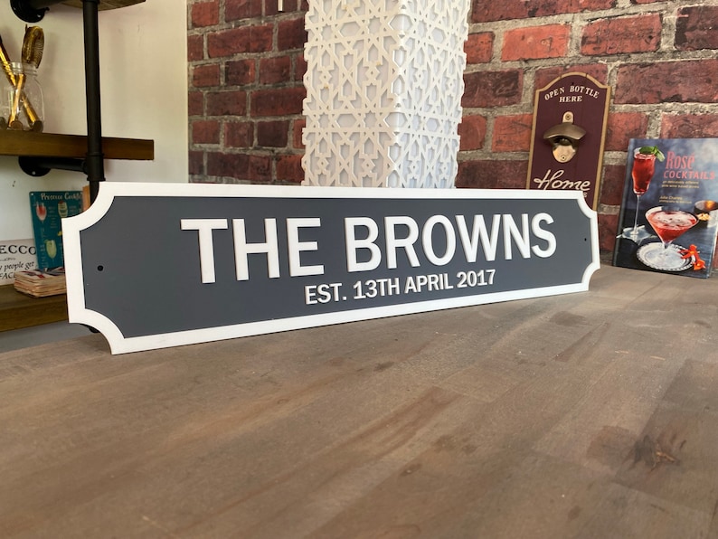 May include: A gray and white sign with the text "THE BROWNS EST. 13TH APRIL 2017". The sign has a white border and is displayed on a wooden surface. The background includes a brick wall and decorative items.