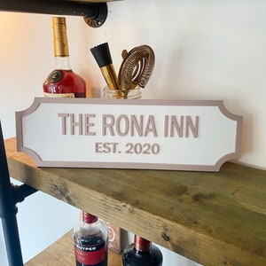 Może przedstawiać: Drewniana półka z tabliczką z napisem "The Rona Inn Est. 2020". Tabliczka jest różowa z białymi literami i ma zaokrąglony górny brzeg. Na półce pod tabliczką stoją dwie butelki alkoholu.