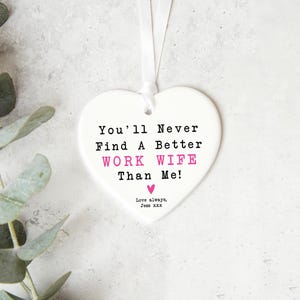 May include: A white heart-shaped ornament with the text "You'll Never Find A Better WORK WIFE Than Me!" in black and pink. A white ribbon is attached for hanging. The words "Love always, Jess XXX" are at the bottom.