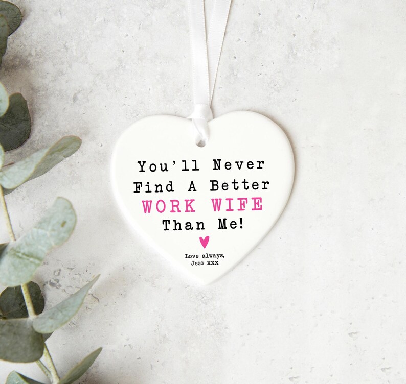 May include: A white heart-shaped ornament with the text "You'll Never Find A Better WORK WIFE Than Me!" in black and pink. A white ribbon is attached for hanging. The words "Love always, Jess XXX" are at the bottom.