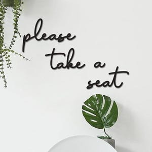 a wall with a sign that says "Please take a seat" in a cursive font. There is also a potted plant and a small green leaf decoration on the wall.