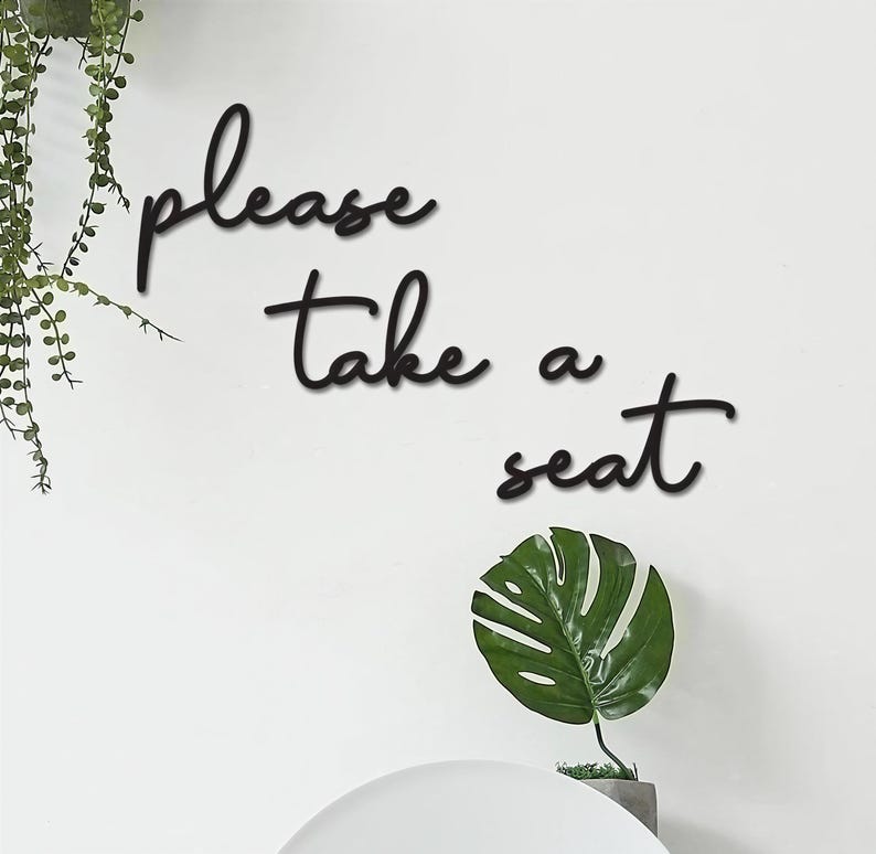 a wall with a sign that says "Please take a seat" in a cursive font. There is also a potted plant and a small green leaf decoration on the wall.