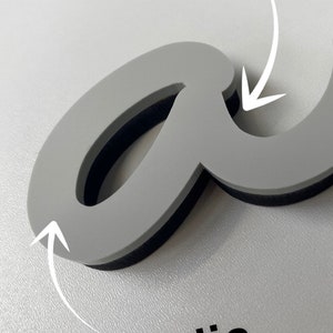 May include: A gray letter "a" made of acrylic with a wood backing. The letter is in a cursive style.