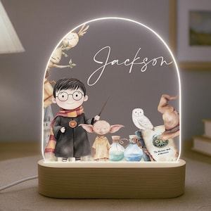 May include: A white and brown night light with a clear acrylic panel featuring a Harry Potter themed illustration. The illustration includes Harry Potter, Hedwig the owl, a house elf, and a book. The name "Jackson" is written in cursive on the panel.