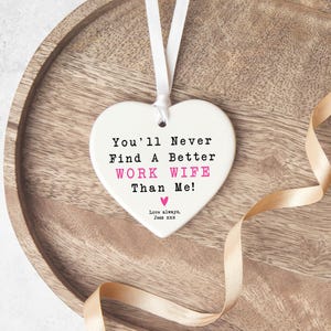 May include: A white heart-shaped ornament with a white ribbon for hanging. The text reads "You'll Never Find A Better WORK WIFE Than Me!" in black and pink. A gold ribbon and wooden tray are in the background.