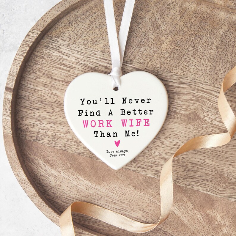 May include: A white heart-shaped ornament with a white ribbon for hanging. The text reads "You'll Never Find A Better WORK WIFE Than Me!" in black and pink. A gold ribbon and wooden tray are in the background.