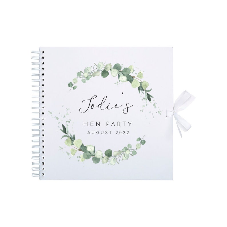 May include: A white spiral bound guest book with a green eucalyptus wreath design. The book has a white ribbon and the text "Sodie's Hen Party August 2022".