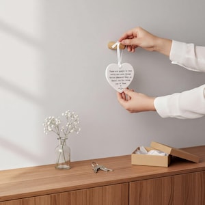 A person's hands holding a heart-shaped ornament with a message, placed on a wooden surface next to a small vase with white flowers and a set of keys.