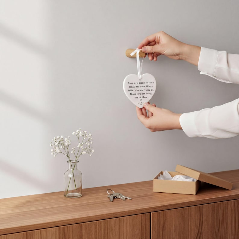 A person's hands holding a heart-shaped ornament with a message, placed on a wooden surface next to a small vase with white flowers and a set of keys.