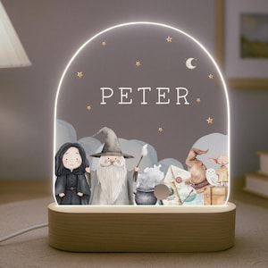 Puede incluir: Una luz nocturna de madera con un panel de acrílico transparente que presenta una ilustración caprichosa de un mago, una bruja, un búho y un caldero. El panel está iluminado con luces LED blancas cálidas. El nombre "PETER" está impreso en el panel.