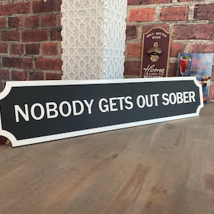 May include: A rectangular black and white sign with the text "NOBODY GETS OUT SOBER". The sign has a white border and is displayed on a wooden surface. The sign is a decorative item.