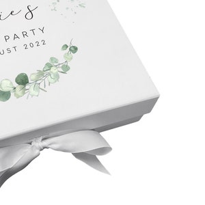 Pode incluir: Caixa de presente branca com uma fita branca amarrada em um la&ccedil;o. A caixa tem um design de coroa de eucalipto verde e o texto "Lie's Party August 2022".