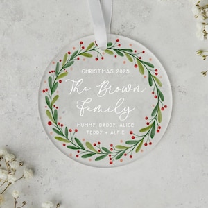 May include: A clear, round Christmas ornament with a white ribbon. The ornament features a green and red wreath design around the edge, with the words "Christmas 2025" and "The Brown Family" in white script. The names "Mummy, Daddy, Alice, Teddy + Alfie" are also included.
