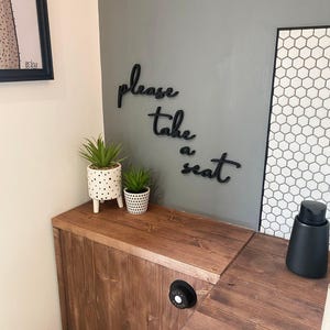 a wooden bench with a sign that says "Please take a seat" on it, along with two potted plants and a black object on the bench.