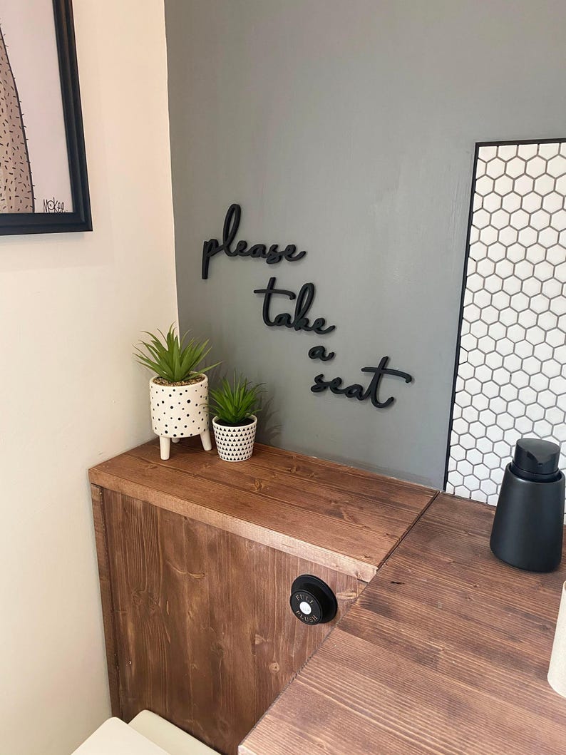a wooden bench with a sign that says "Please take a seat" on it, along with two potted plants and a black object on the bench.