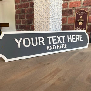 May include: A rectangular sign with a gray background and white trim. The sign reads "YOUR TEXT HERE" and "AND HERE" in white, bold letters. The sign is displayed on a wooden surface.
