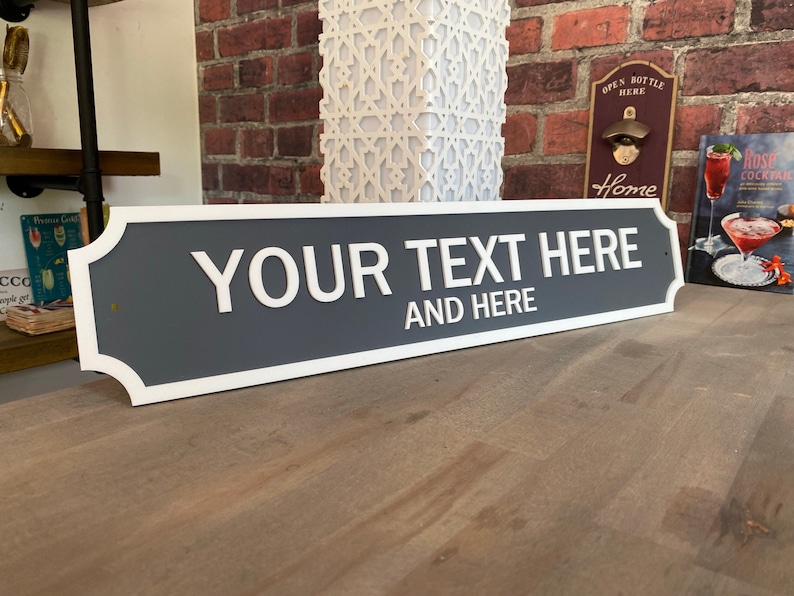 May include: A rectangular sign with a gray background and white trim. The sign reads "YOUR TEXT HERE" and "AND HERE" in white, bold letters. The sign is displayed on a wooden surface.