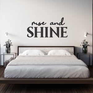 May include: A white bedroom with a bed and two nightstands. The wall behind the bed has a black vinyl decal that reads "rise and shine".