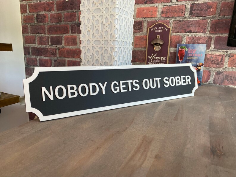 May include: A black and white sign with rounded edges and a white border. The sign reads "Nobody Gets Out Sober".