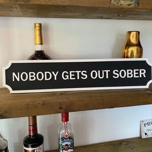May include: Black and white sign with rounded corners and white trim that reads "Nobody Gets Out Sober". The sign is mounted on a wooden shelf with bottles of alcohol on either side.