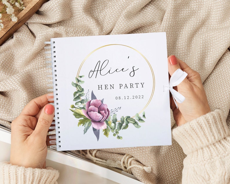 May include: A white spiral bound book with a gold circle frame and floral design. The text "Alice's Hen Party 08.12.2022" is printed on the cover.