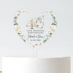 Può includere: Un cake topper a forma di cuore trasparente, con la scritta "40TH WEDDING ANNIVERSARY", i nomi "Deborah & Chris" e la data "01.08.1986". Il design include elementi floreali e dorati.