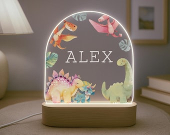 Könnte beinhalten: Ein Nachtlicht aus Holz mit einer durchsichtigen Acrylscheibe mit Dinosaurier-Motiv. Die Scheibe zeigt ein Aquarelldesign mit Dinosauriern und Blättern. Der Name "ALEX" ist in Weiß auf die Scheibe gedruckt.