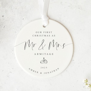 Może przedstawiać: Okrągła, biała ceramiczna ozdoba świąteczna z czarnym napisem. Tekst brzmi "OUR FIRST CHRISTMAS AS Mr & Mrs ARMITAGE", z imionami pary, rokiem 2025 i wstążką do zawieszenia. Minimalistyczny design.
