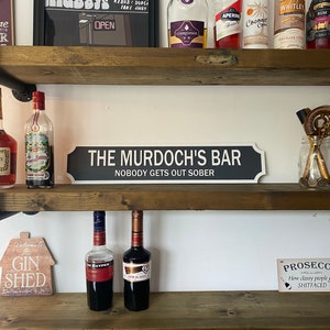 May include: A black and white sign that reads "THE MURDOCH'S BAR NOBODY GETS OUT SOBER" is displayed on a wooden shelf. The shelf also holds various bottles of liquor and bar accessories, creating a home bar theme.