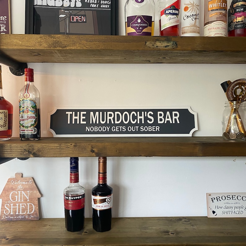 May include: A black and white sign that reads "THE MURDOCH'S BAR NOBODY GETS OUT SOBER" is displayed on a wooden shelf. The shelf also holds various bottles of liquor and bar accessories, creating a home bar theme.