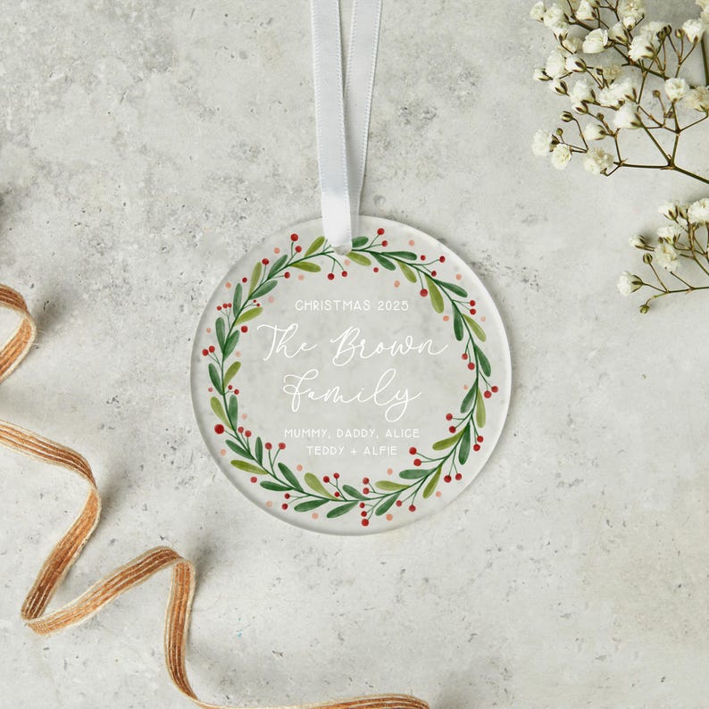 May include: A clear, round Christmas ornament with a white ribbon. The ornament features a green wreath with red berries and the text "The Brown Family" and "Christmas 2025".
