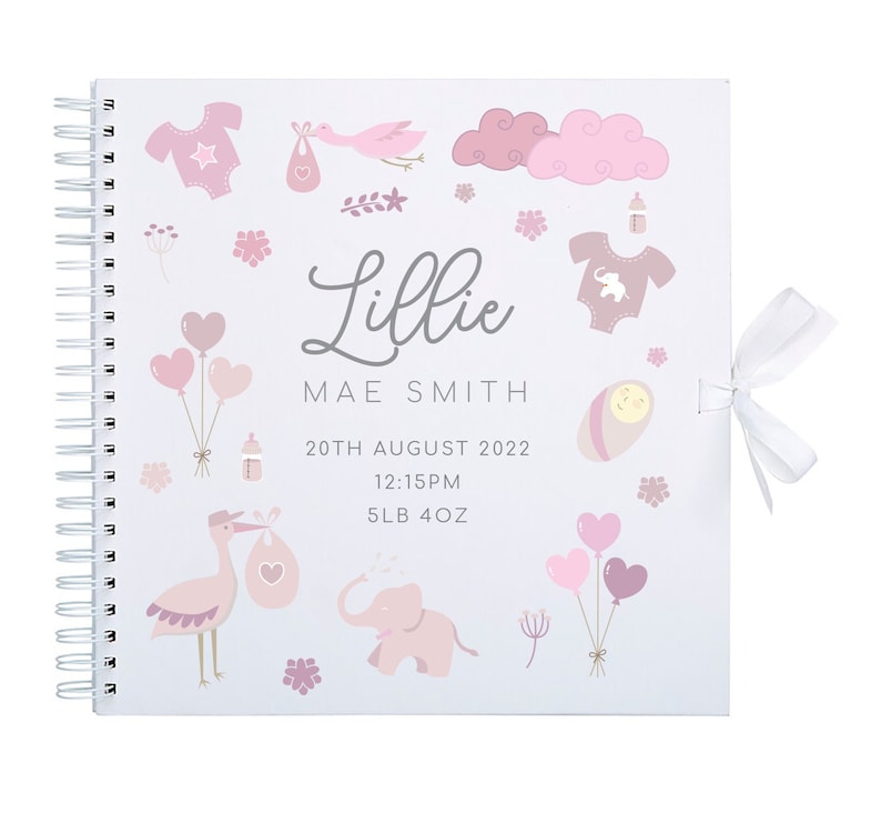 May include: A white baby memory book with a pink ribbon. The cover features illustrations of a baby, a stork, a cloud, balloons, and an elephant. The text on the cover reads "Lillie Mae Smith, 20th August 2022, 12:15PM, 5LB 4OZ."