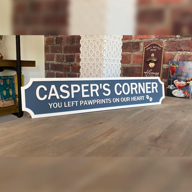 May include: A blue and white sign that reads "CASPER'S CORNER" with the text "YOU LEFT PAWPRINTS ON OUR HEART" and a paw print graphic. The sign is rectangular with a decorative border.