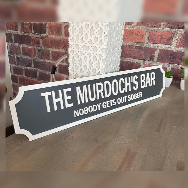 May include: A rectangular sign with a dark gray center and white trim. The sign reads "THE MURDOCH'S BAR" above "NOBODY GETS OUT SOBER" in white lettering. The sign is displayed on a wooden surface.