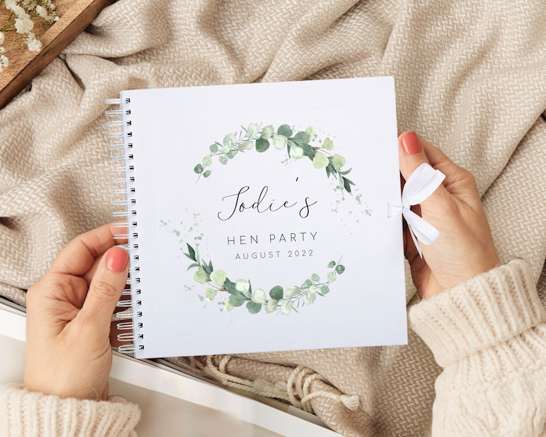 May include: A white spiral bound journal with a green eucalyptus wreath design. The text "Jodie's Hen Party August 2022" is printed on the front cover.