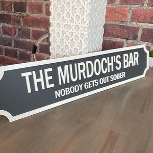 May include: A rectangular sign with the text "THE MURDOCH'S BAR" and "NOBODY GETS OUT SOBER" in white lettering. The sign has a dark gray center with a white border and is set against a brick wall.