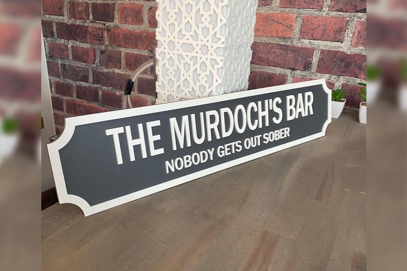 May include: A rectangular sign with the text "THE MURDOCH'S BAR" and "NOBODY GETS OUT SOBER" in white lettering. The sign has a dark gray center with a white border and is set against a brick wall.