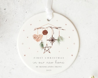 May include: A round, white Christmas ornament with a watercolor design. The ornament features a branch with hanging decorations, including a heart, star, and greenery. Text reads "FIRST CHRISTMAS in our new home" and address and year.