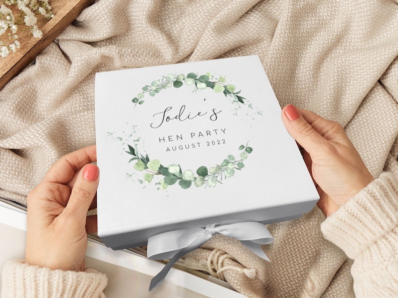 Pode incluir: Caixa de presente branca com uma fita cinza. A caixa tem um design de coroa de eucalipto verde com o texto "Jodie's Hen Party August 2022".
