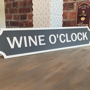 May include: Grey and white sign with rounded edges that reads "Wine O'Clock".